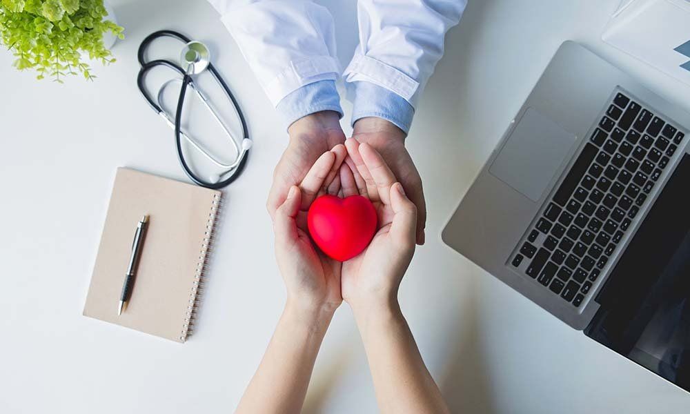 A doctor in a white coat holds a red heart symbol with a patient's hands above a desk with a stethoscope and laptop.