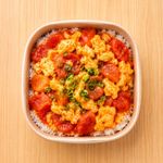 Square bowl of rice topped with scrambled eggs, sliced red sausage, and green garnish on a wooden table