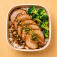 Sliced glazed chicken with broccoli over rice in a square takeout container