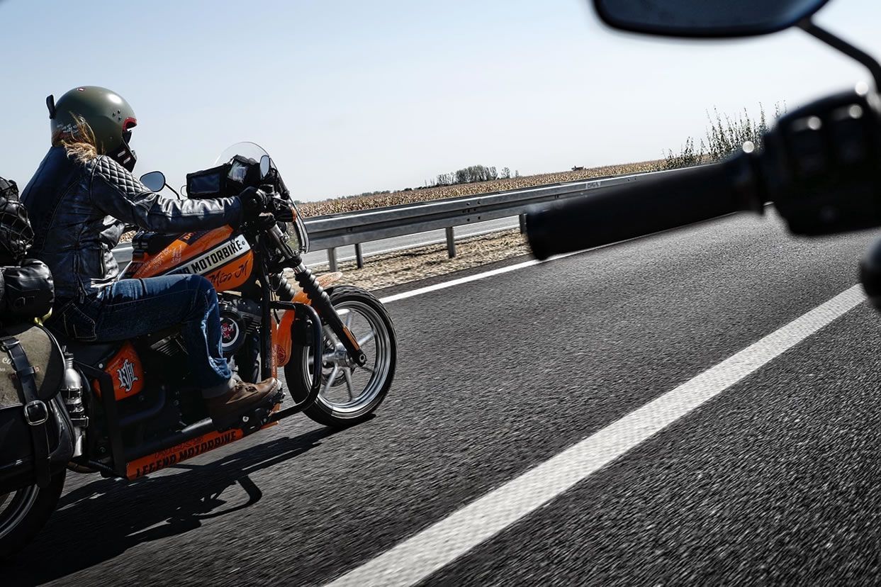 Un motard sur une moto orange circule sur une route goudronnée, à côté du guidon d'une autre moto. Journée ensoleillée.