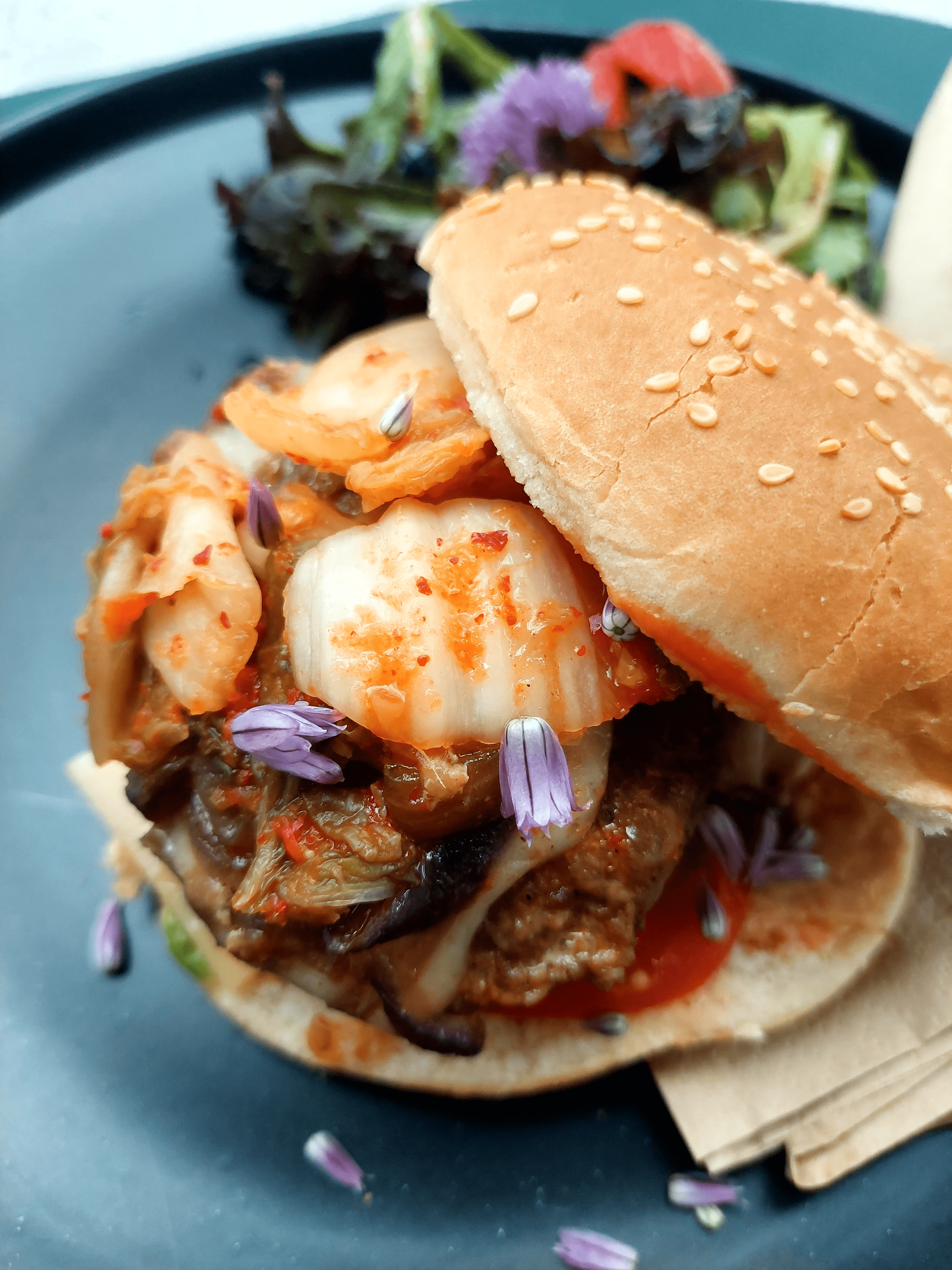 Burger au kimchi avec pain au sésame, servi avec de la salade sur une assiette bleue.