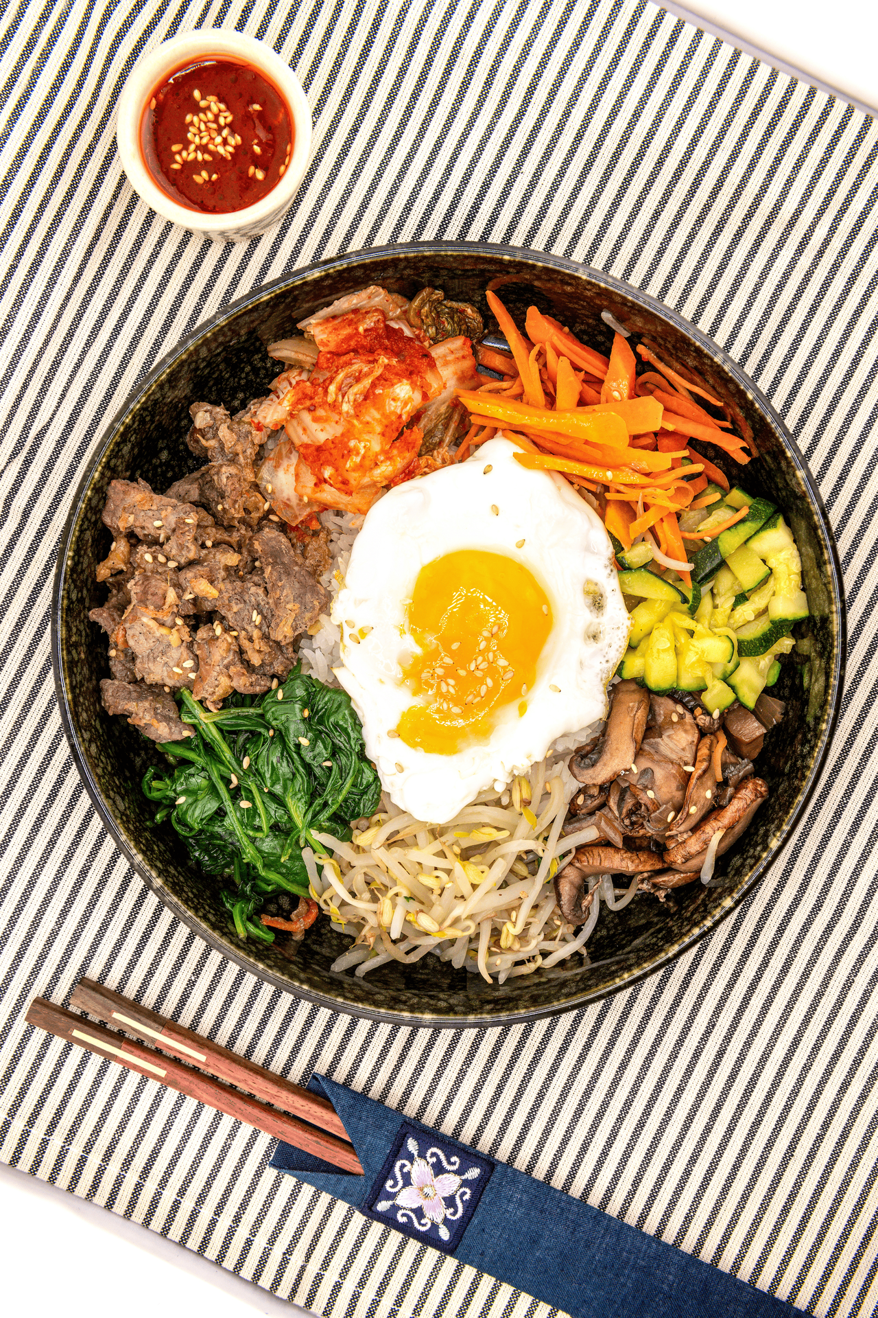 Bol de bibimbap avec un assortiment de légumes, de viande et un œuf au plat, à côté d'une sauce et de baguettes.