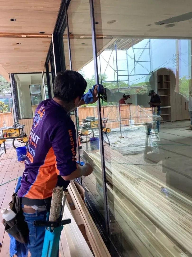Man cleaning large glass window, indoors. He wears a purple and orange work shirt and uses a tool.