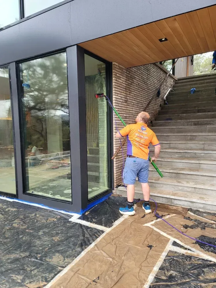 Man in orange shirt cleans exterior window with a pole. Building with black trim and steps.