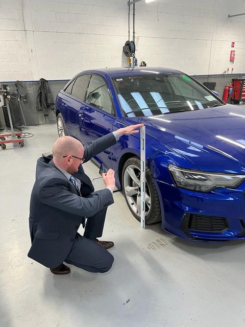 Man in suit measures the front of a blue car in a garage.