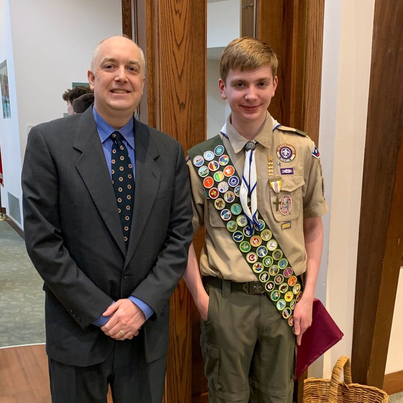 Two people standing indoors, one in a suit and one in a scout uniform with merit badges.