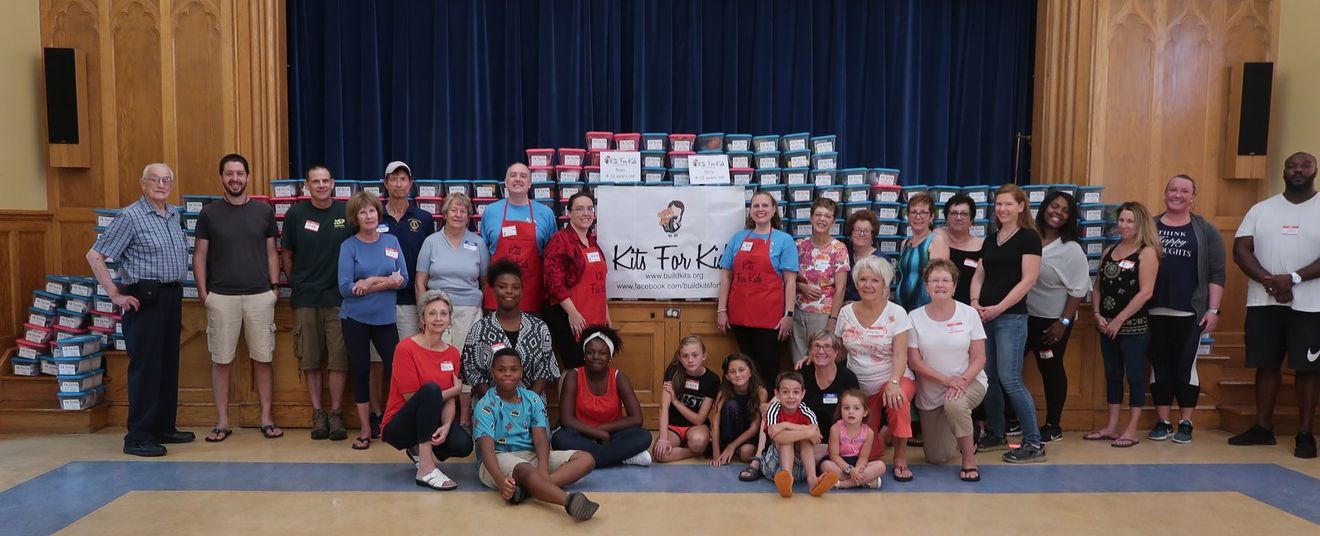 Large group posing on a stage with blue curtains and a backdrop of sponsor signs, holding donation boxes and gifts