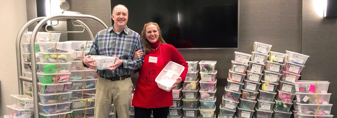 Two people stand beside stacks of colorful container sets in a store aisle, one holding a white box.