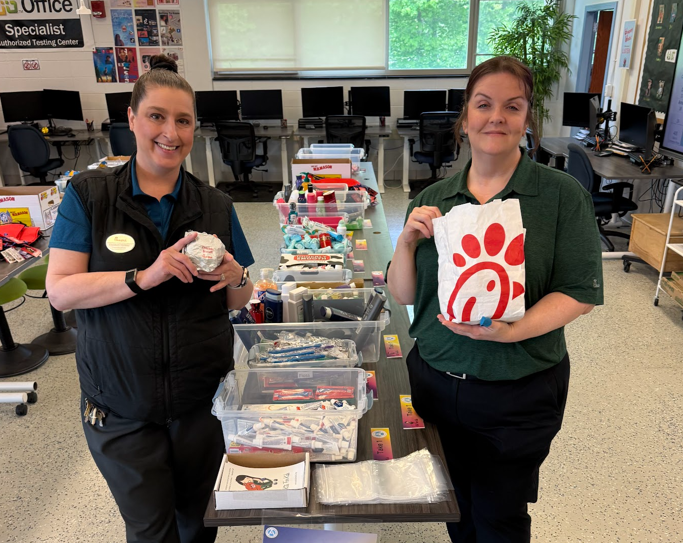 Two staff members stand behind a table of boxed items, one holding a Chick-fil-A bag in a classroom setting.