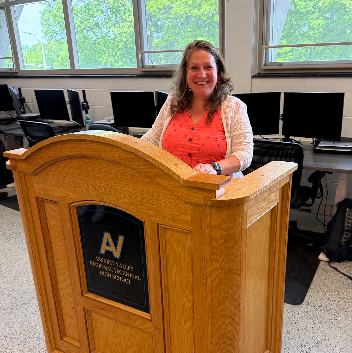 Woman smiling behind a wooden podium in a computer lab or classroom, with monitors and windows behind her