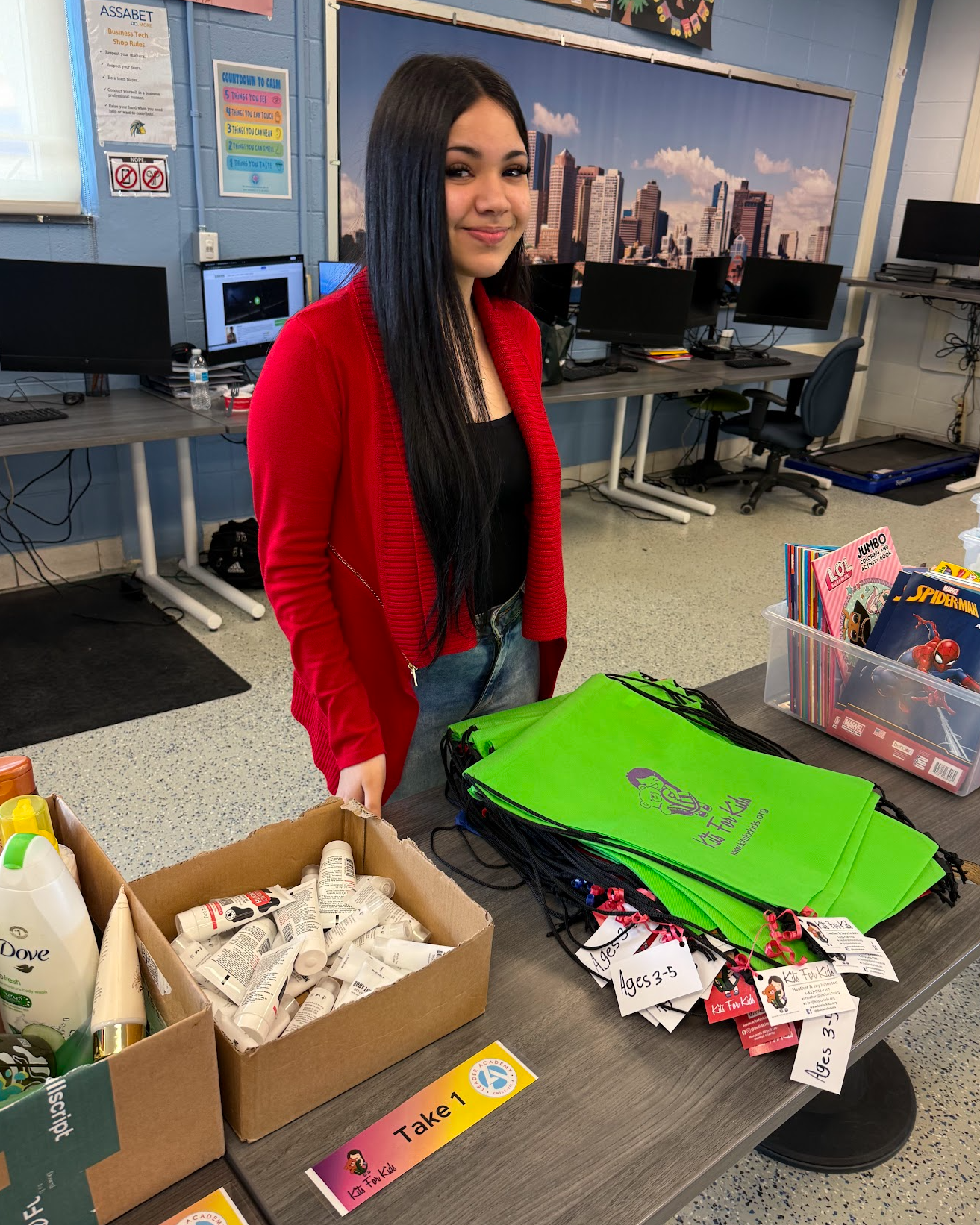 Person standing beside a table with craft supplies in a classroom, wearing a red cardigan.