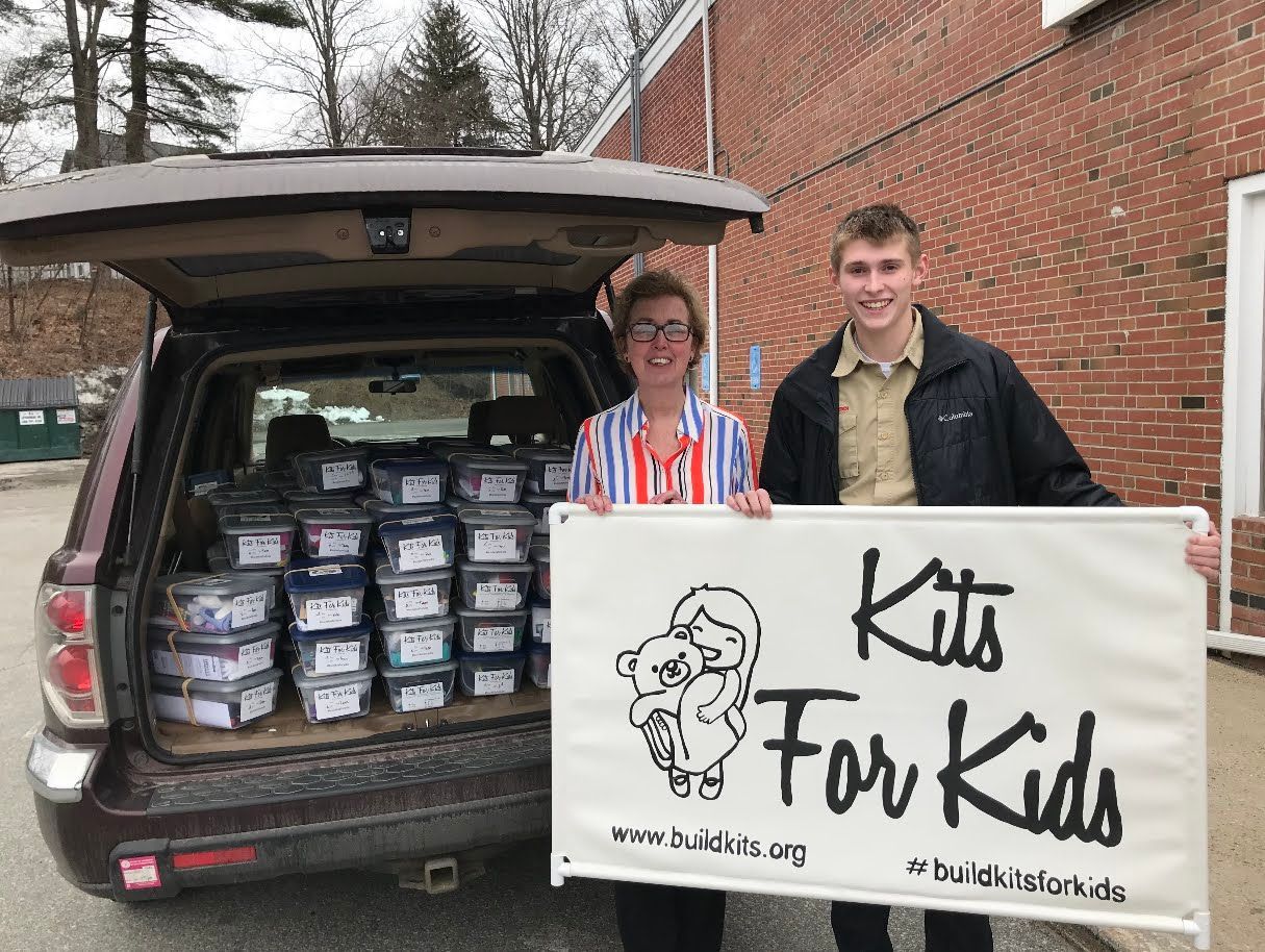 People standing by an SUV loaded with food boxes, holding a “Kids for Kid” sign outside a brick building