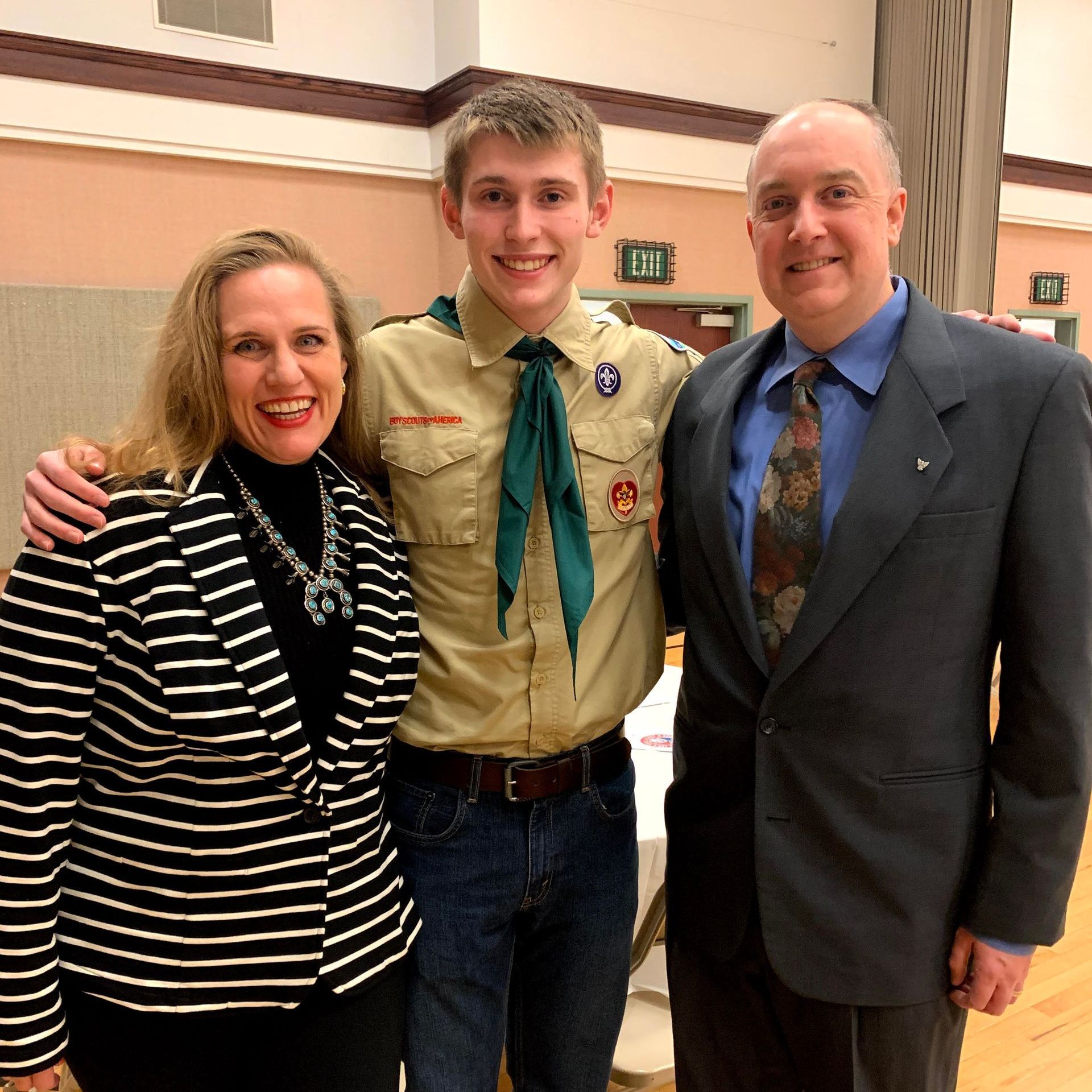 Three people posing indoors, with a scout in uniform between a woman in stripes and a man in a suit.