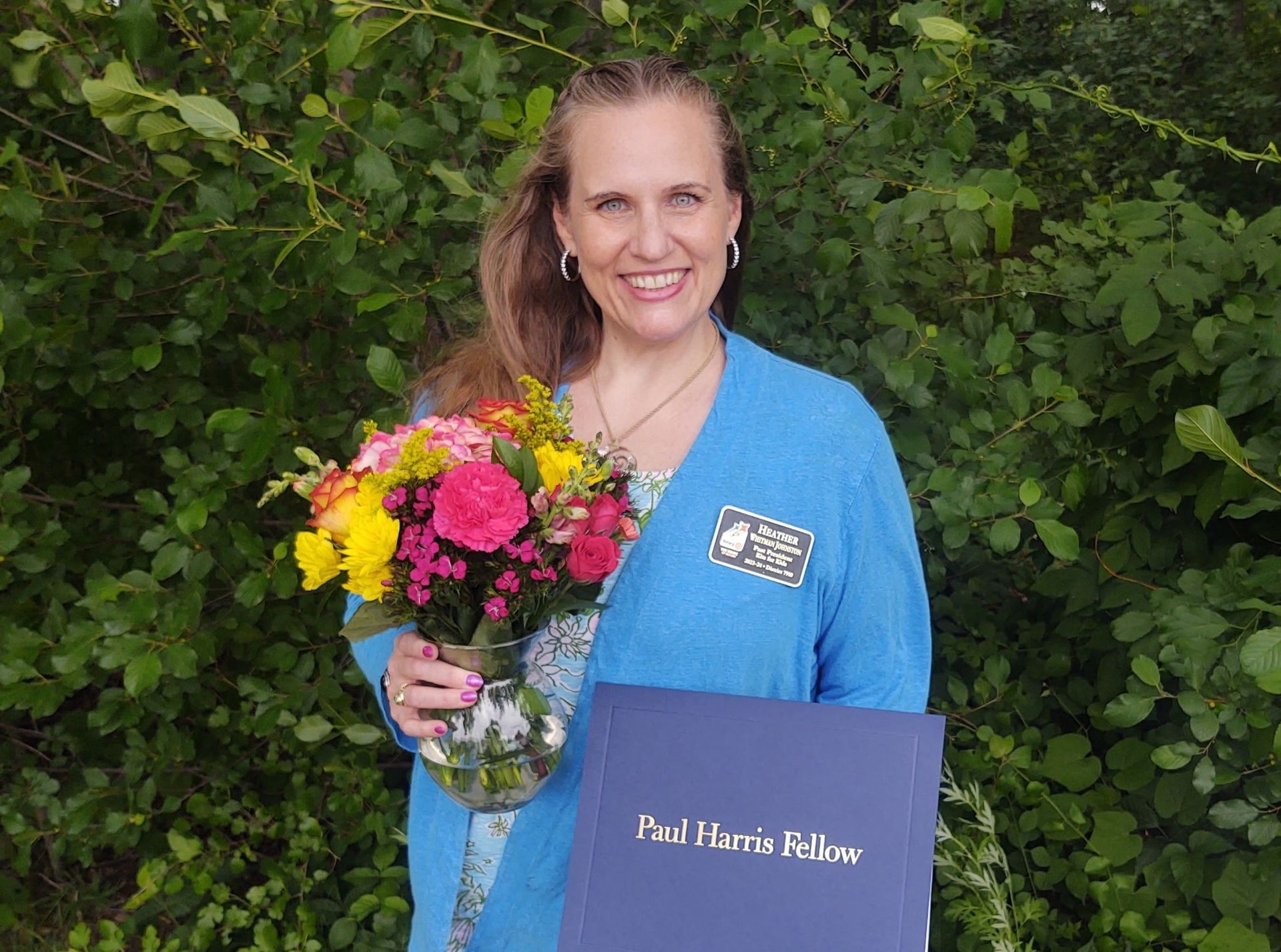 Woman holding a colorful bouquet and a blue diploma folder in front of green foliage