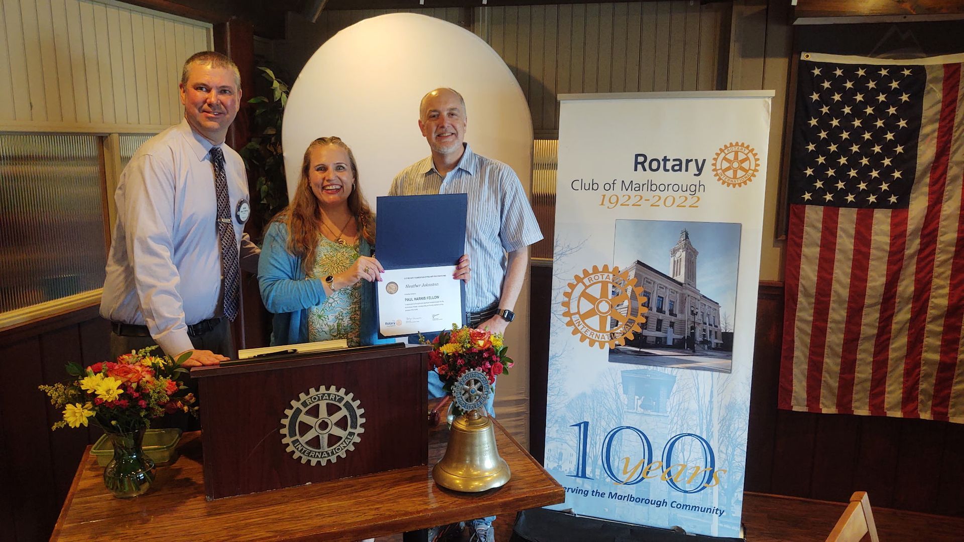Three people pose at a Rotary event, holding a certificate beside a Rotary 100th anniversary banner.