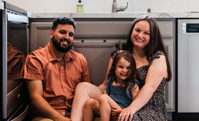 Three people smiling on a couch, with a child seated between two adults in a living room.