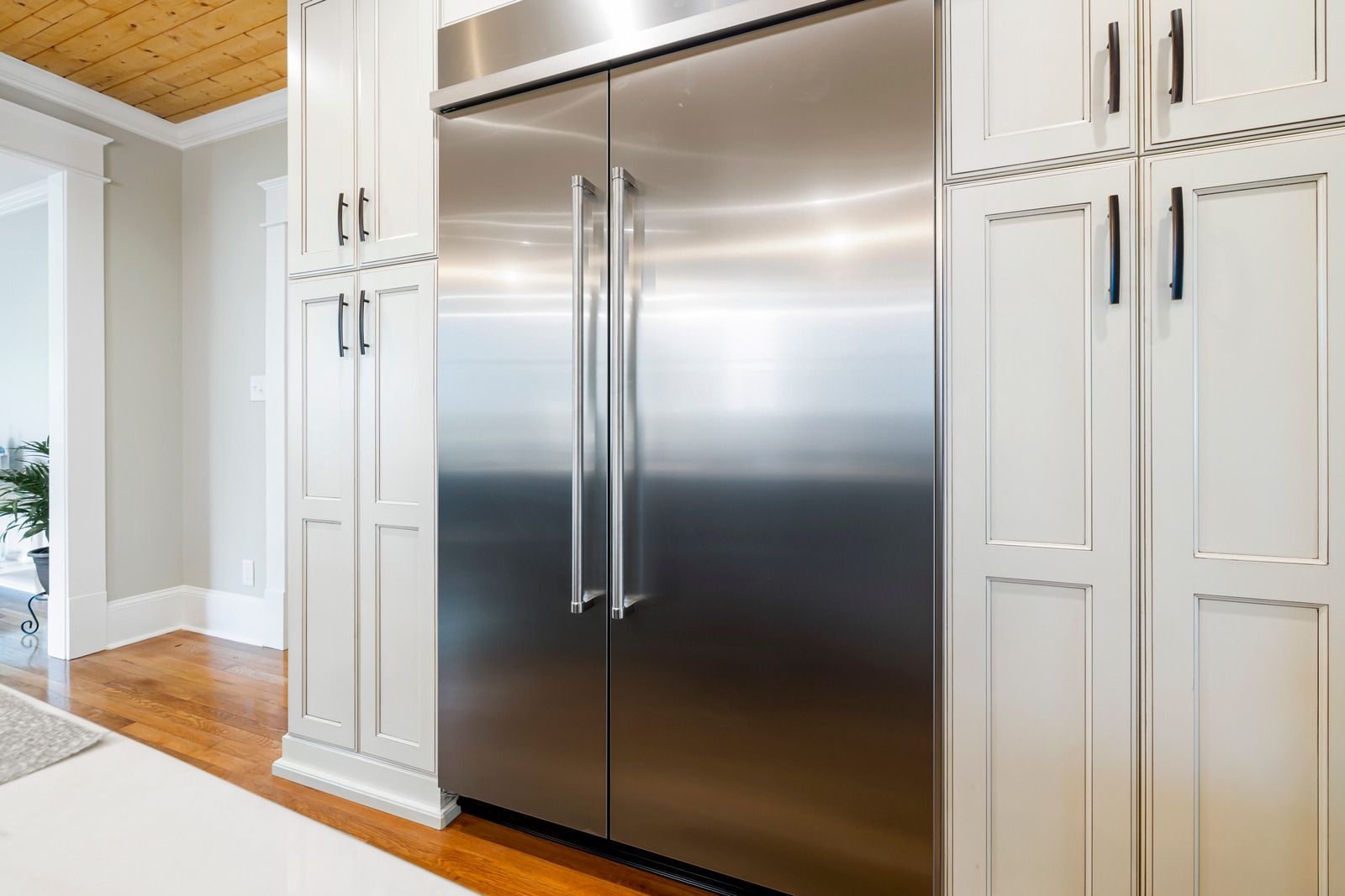 Stainless steel double-door refrigerator built into white kitchen cabinetry with wood ceiling and floors