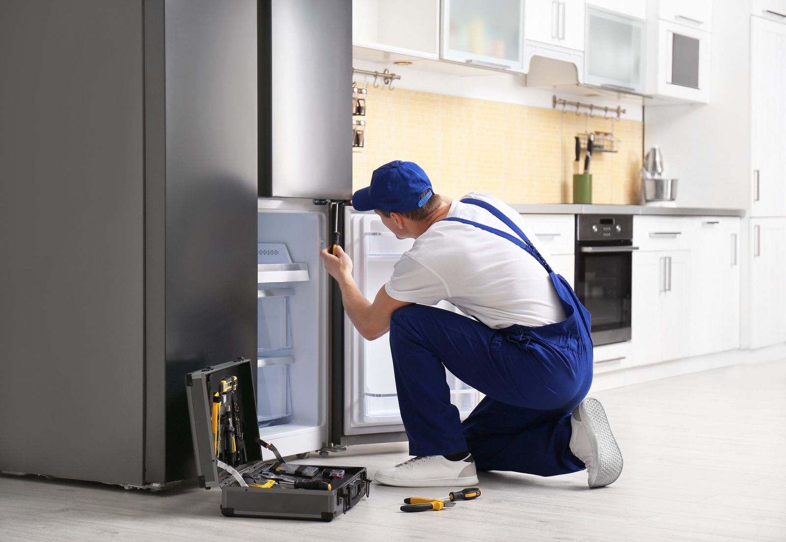 A repairman in blue overalls kneels, fixing a refrigerator in a kitchen with a toolbox open nearby.