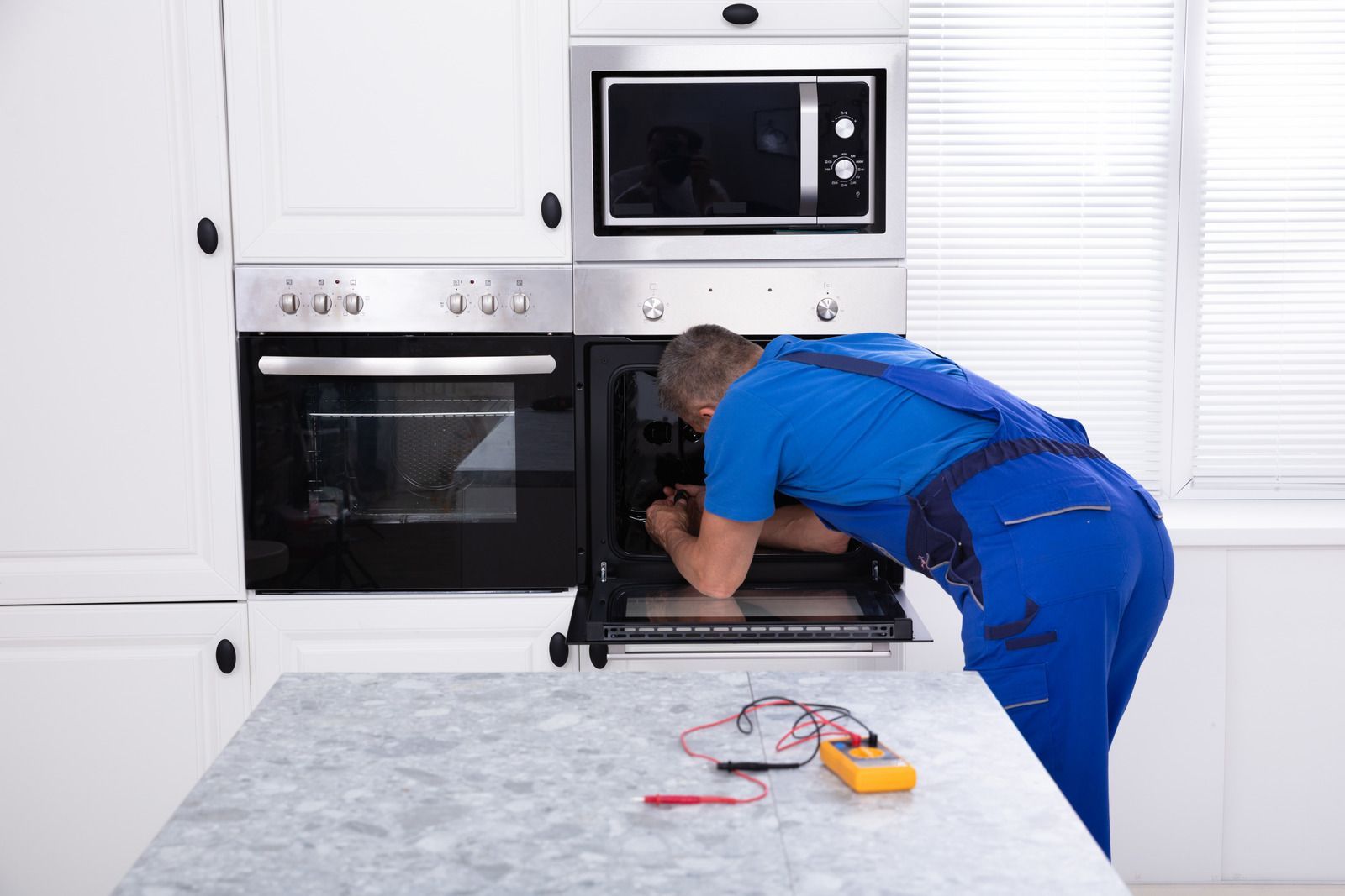 Technician in blue coveralls inspecting an oven in a white kitchen, with tools on the counter.