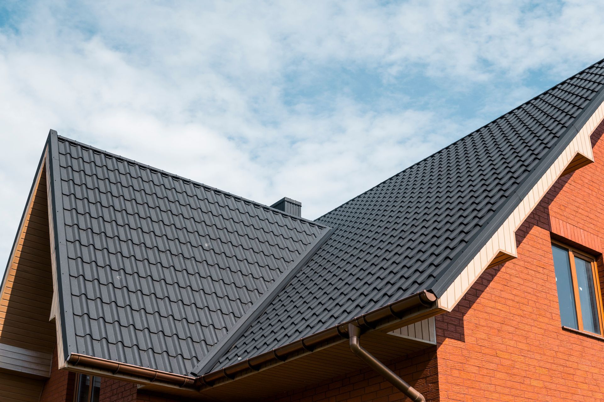 Modern house roof with dark gray tiles and orange brick walls under a blue sky
