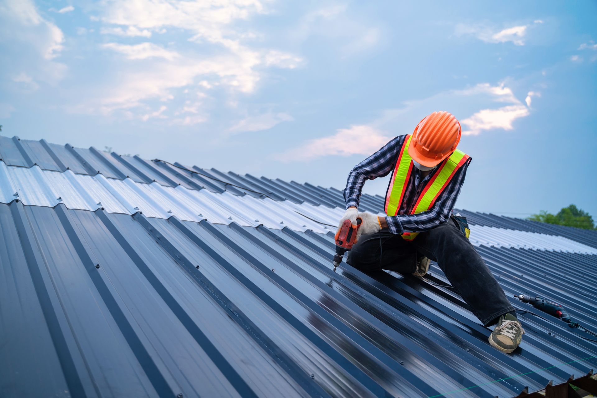 Worker in safety gear installing roofing panels on a metal roof under a blue sky