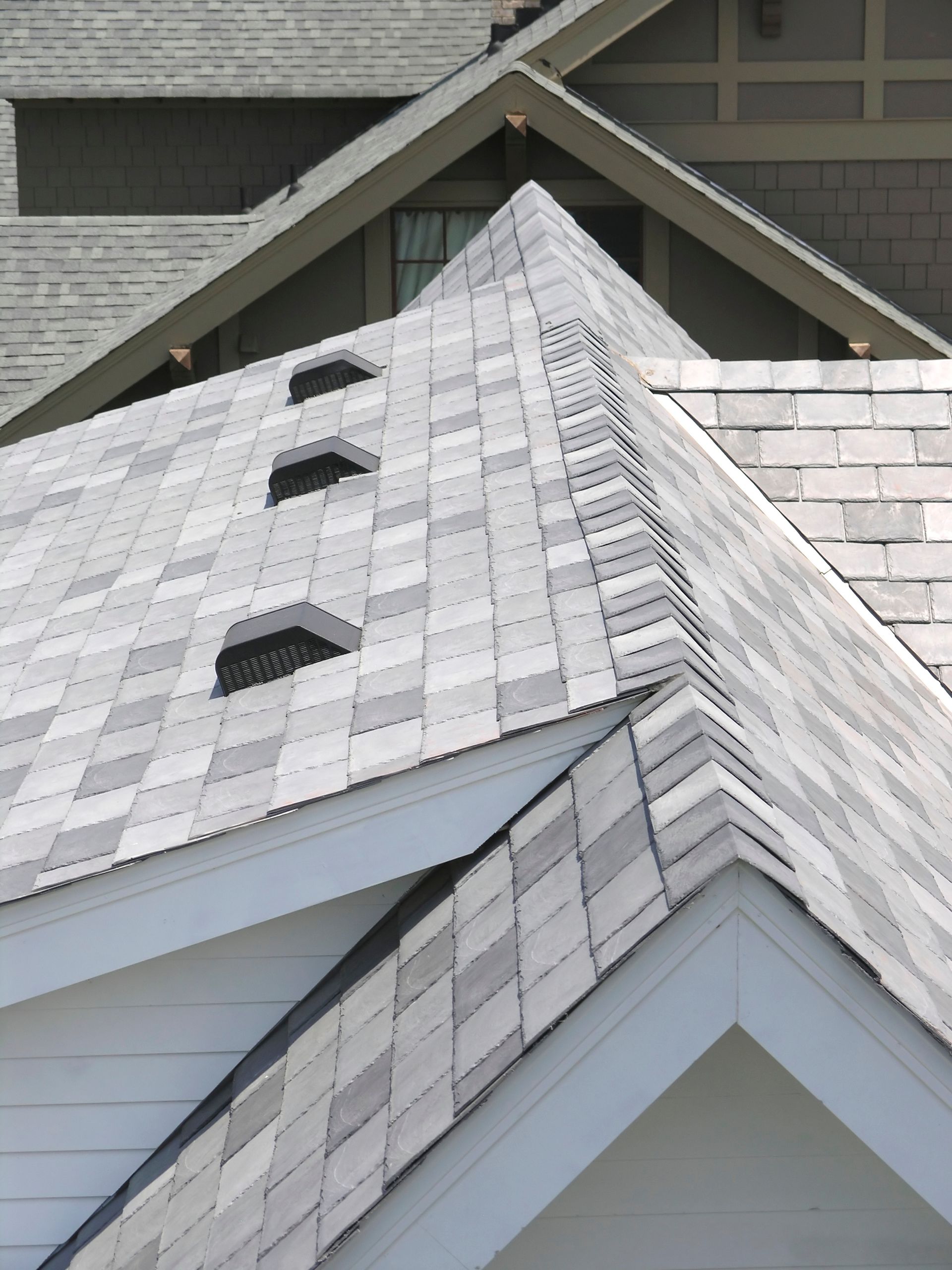 Close-up of a gray shingled roof with several vents and an adjoining roofline against a house exterior