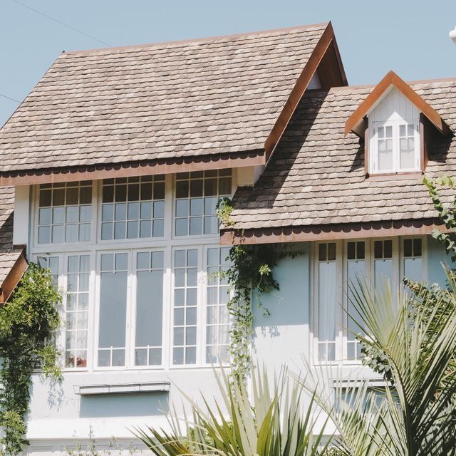 White house with large windows, steep shingled roof, and dormer, framed by tropical plants