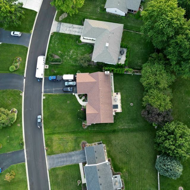 Aerial view of suburban homes, lawns, parked cars, and a curved street surrounded by trees.