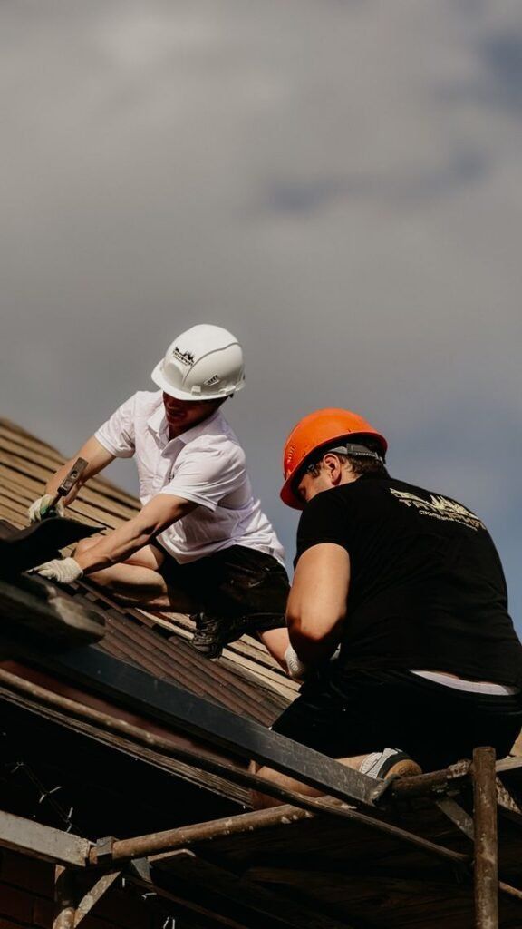 Two workers in hard hats crouch on a roof, inspecting or repairing shingles.