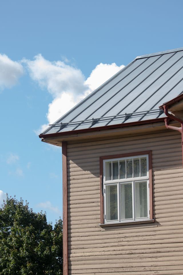 Close-up of a house exterior with beige siding, white-trimmed window, and gray metal roof under a blue sky.
