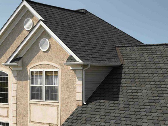 Two-story beige house with dark gray shingled roof and white-trimmed windows under a blue sky