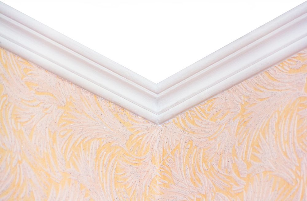 Una modanatura bianca sul soffitto, sopra una carta da parati a motivi color pesca chiaro, forma un dettaglio angolare.