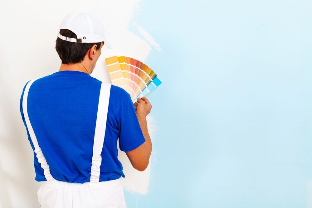 Pittore con camicia blu e tuta bianca che tiene in mano un campionario di colori contro una parete bianca