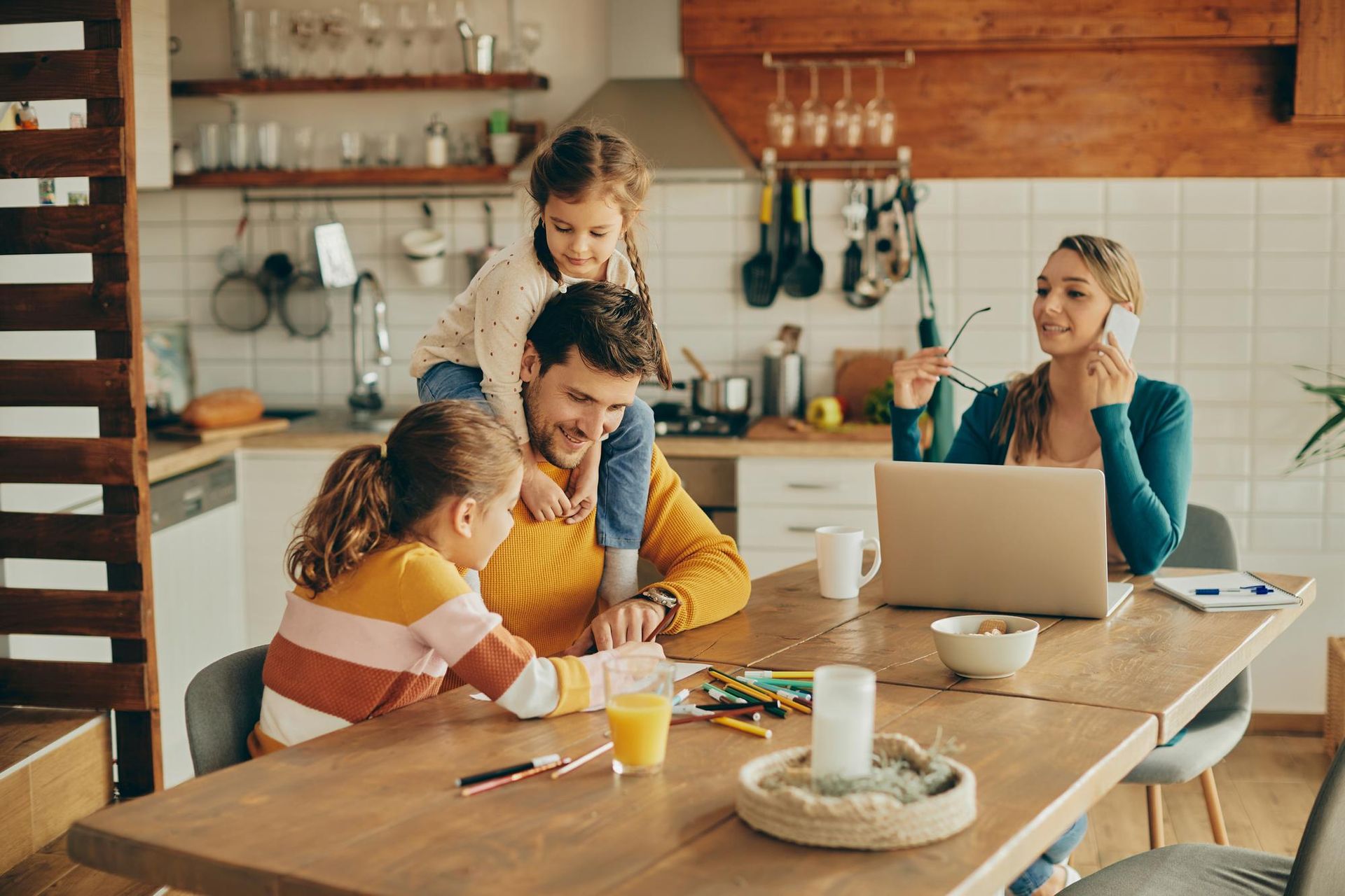 Family gathered around a wooden table in a cozy kitchen, using a laptop and sharing a meal together.
