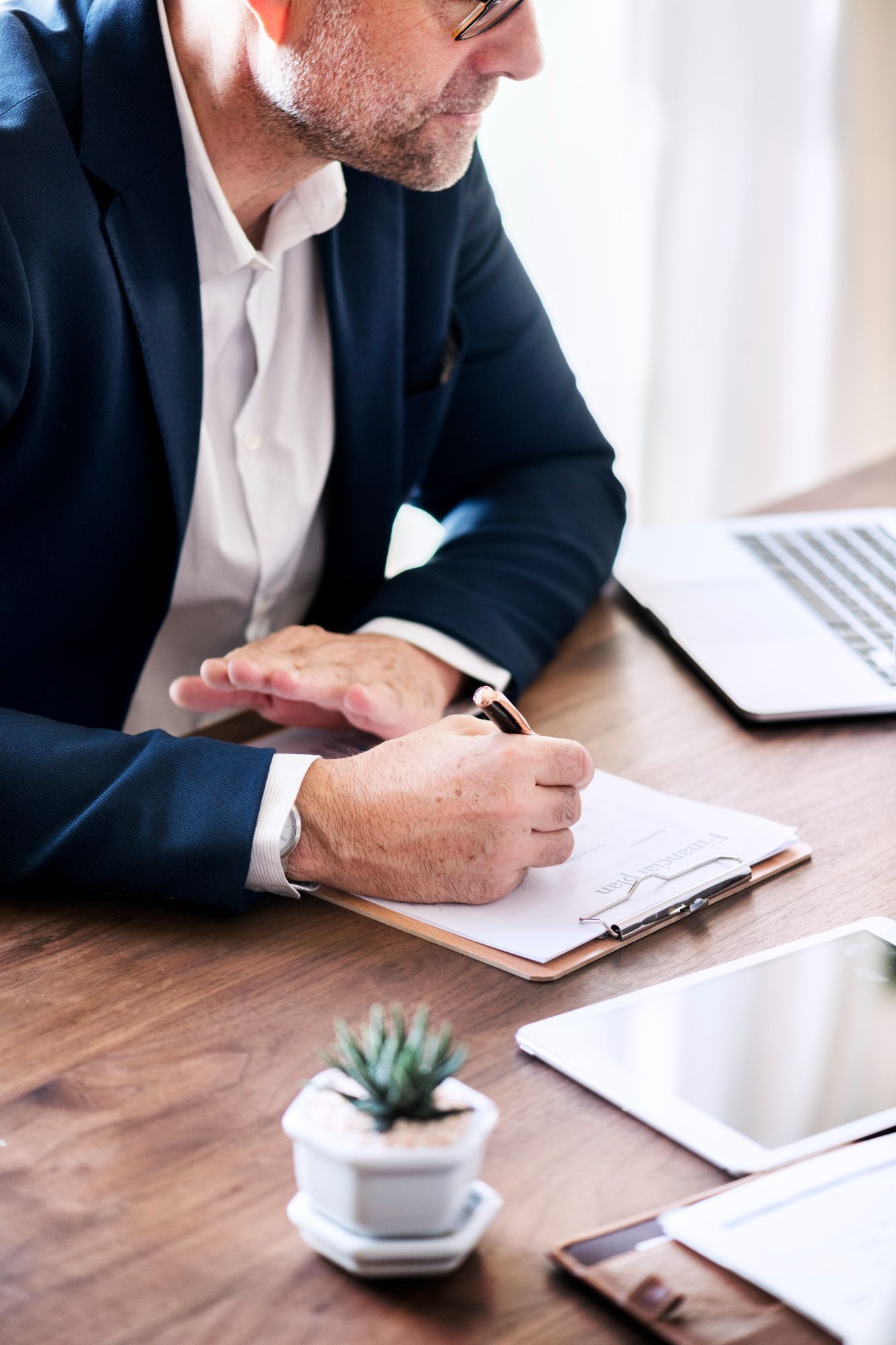 Businessperson writing in a notebook at a desk with a laptop and tablet nearby