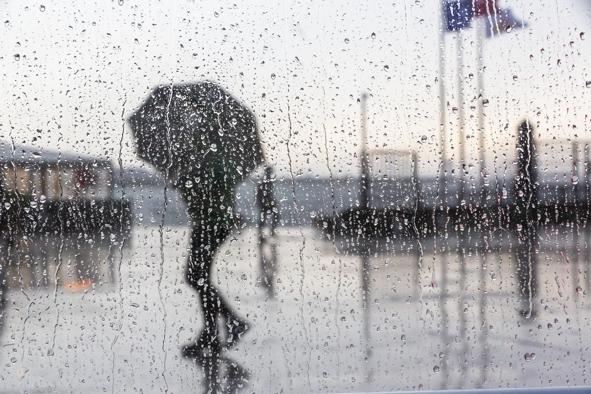 Person with umbrella walking in the rain behind a rain-streaked window.