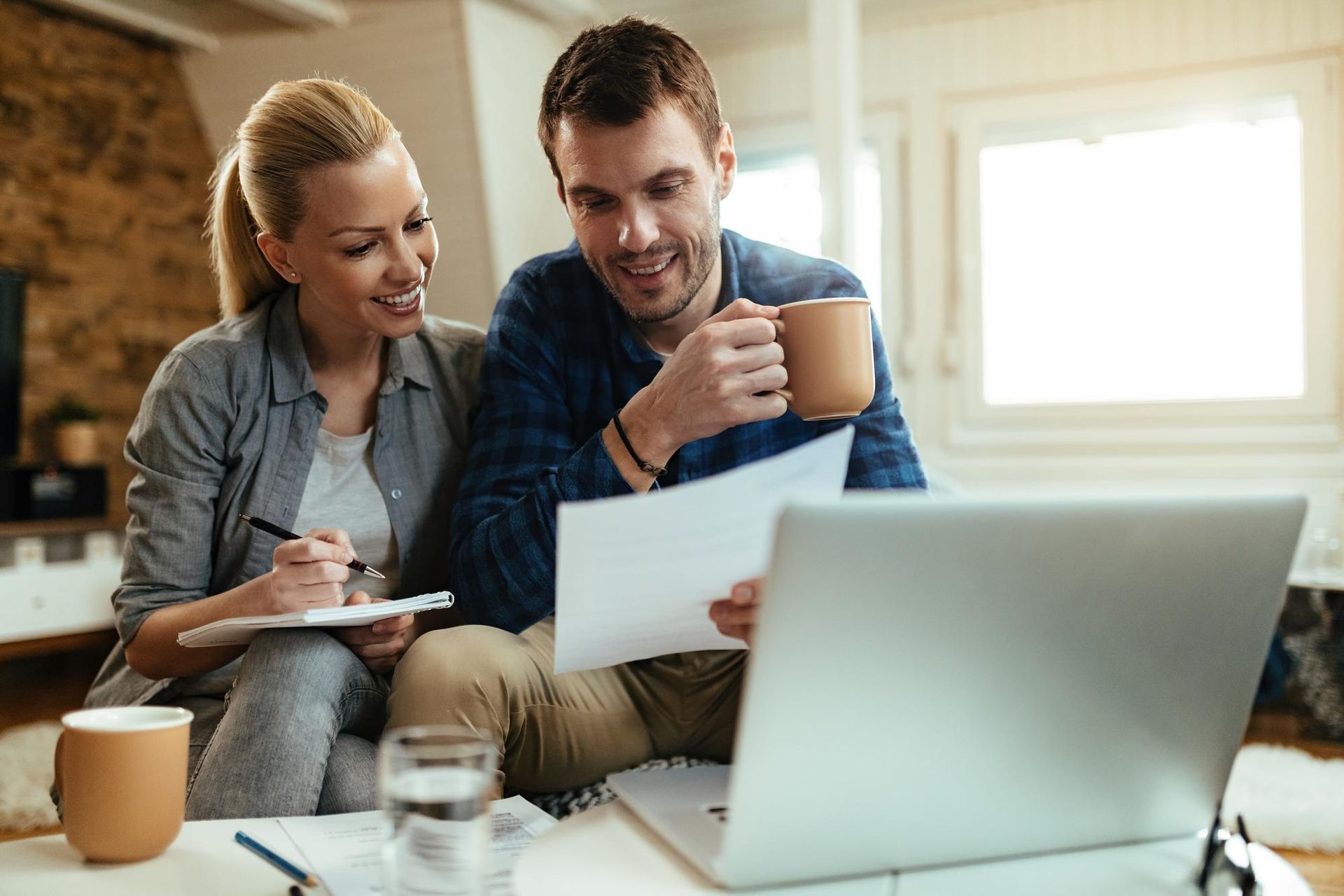 Two people on a couch reviewing papers beside a laptop, holding coffee mugs in a bright room.