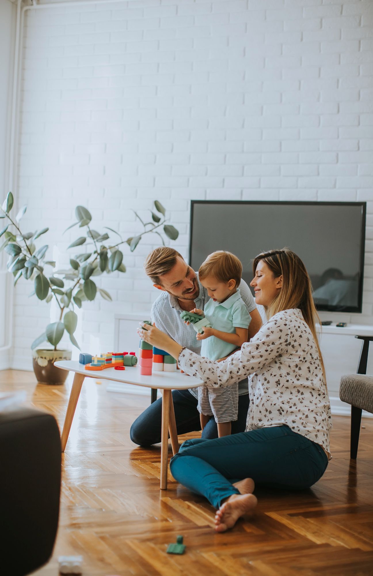 Family playing with a toddler on the floor in a bright living room, with a table and TV nearby
