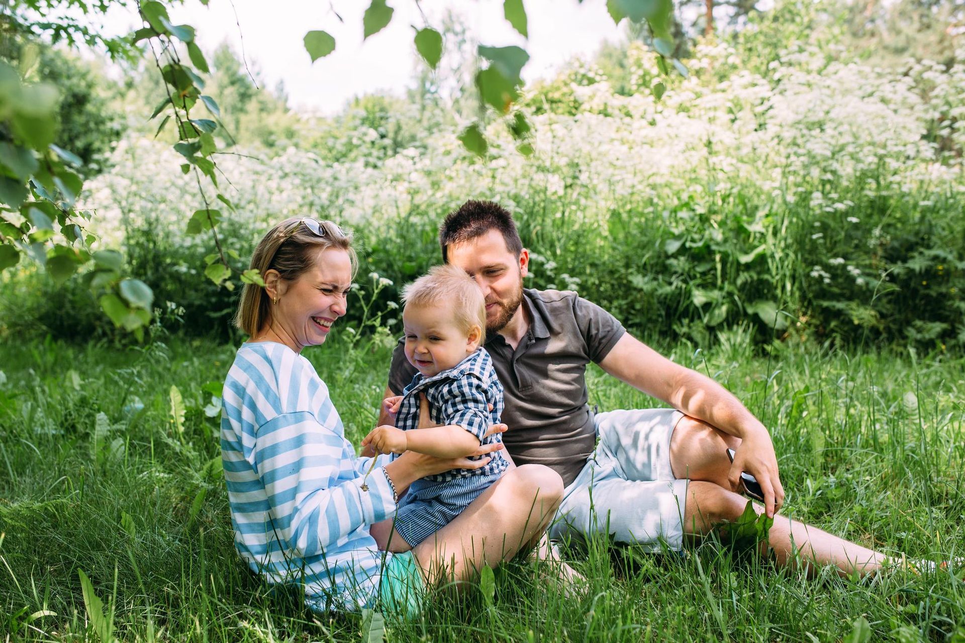 Family sitting on grass in a sunny garden, smiling with a baby among white flowers.