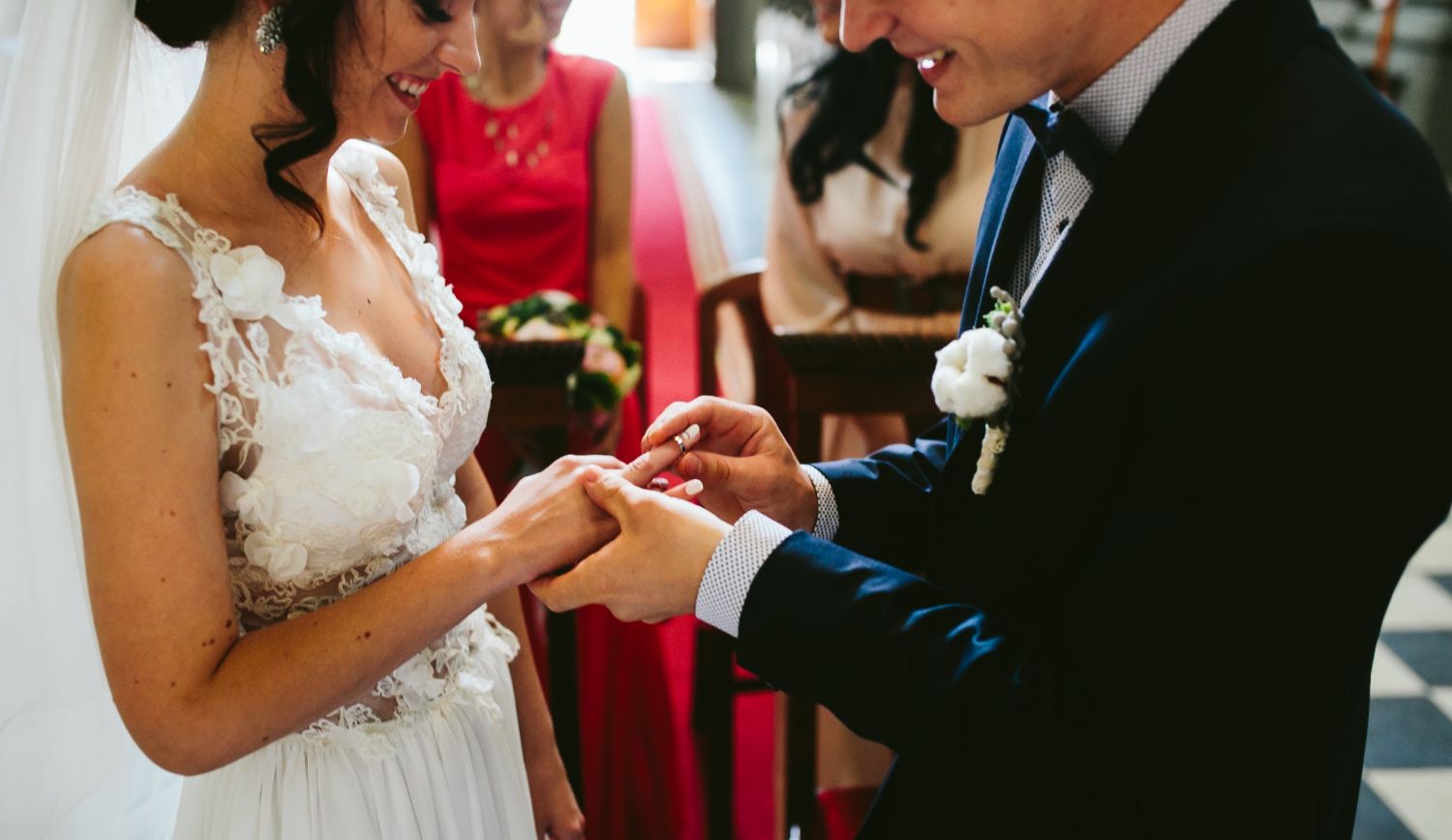Bride and groom exchanging rings during a wedding ceremony, with a red-dressed attendant in the background