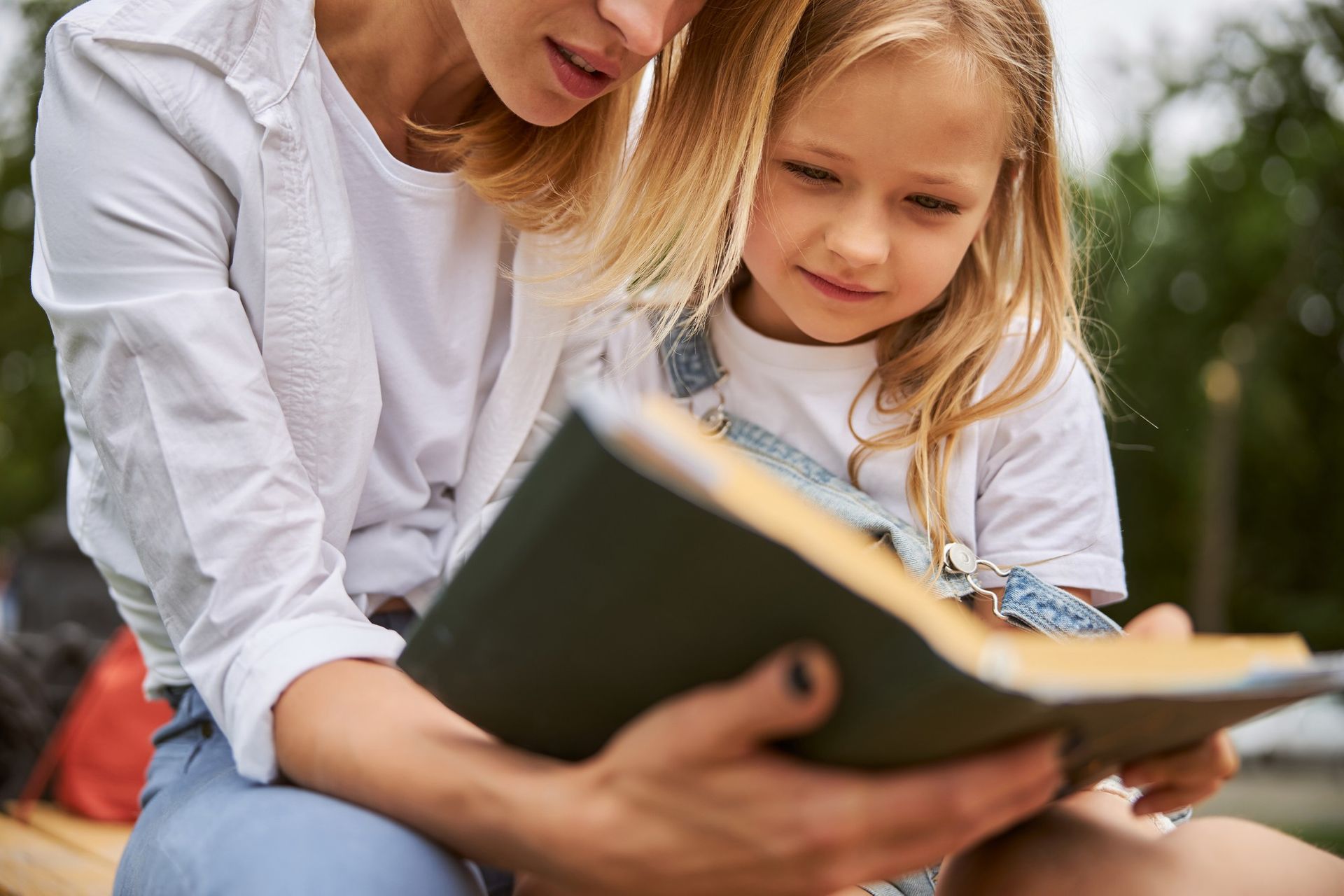 Adult and child reading a book together outdoors, smiling in a sunny park