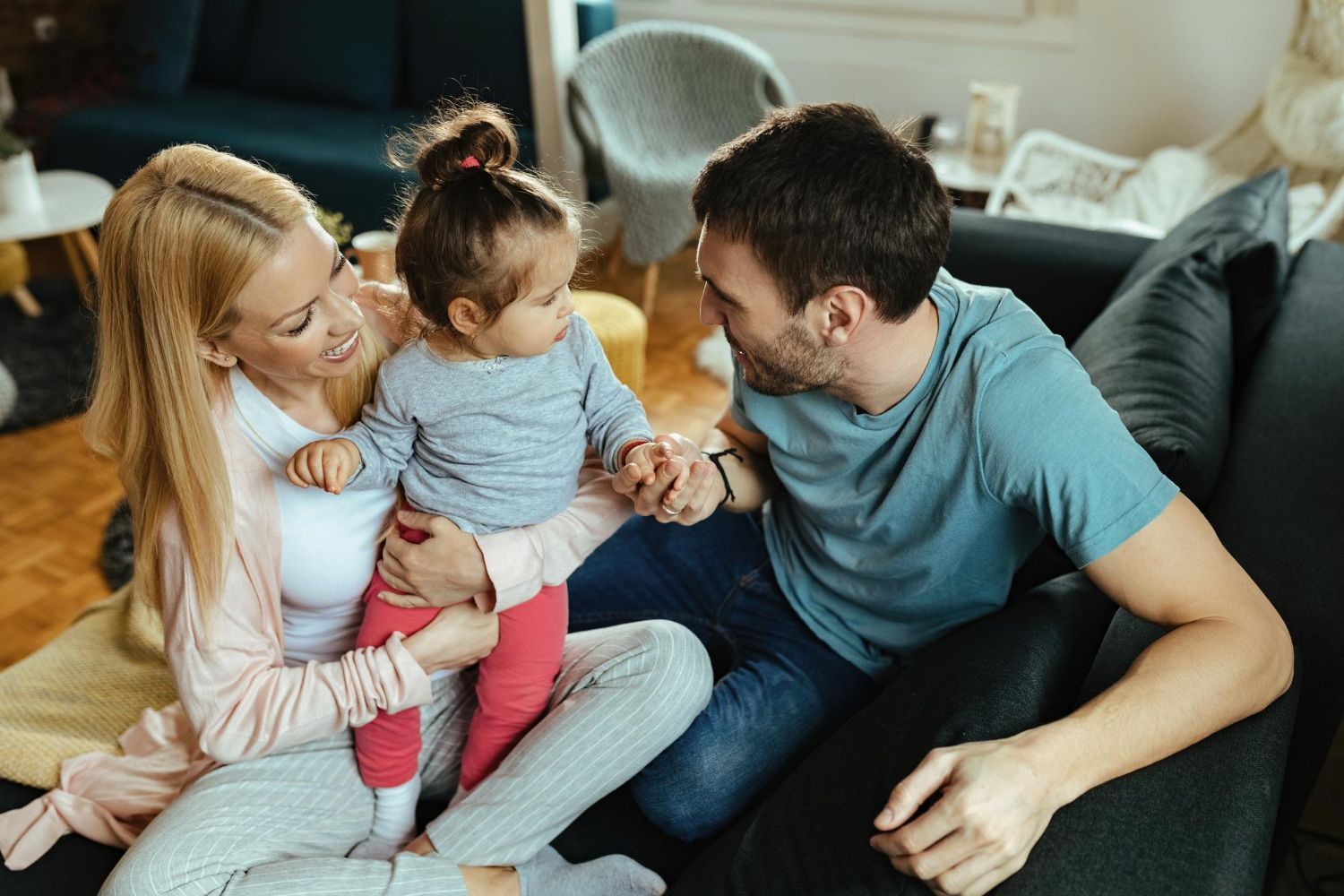 Family sitting on a couch, parents smiling at a toddler in a cozy living room.