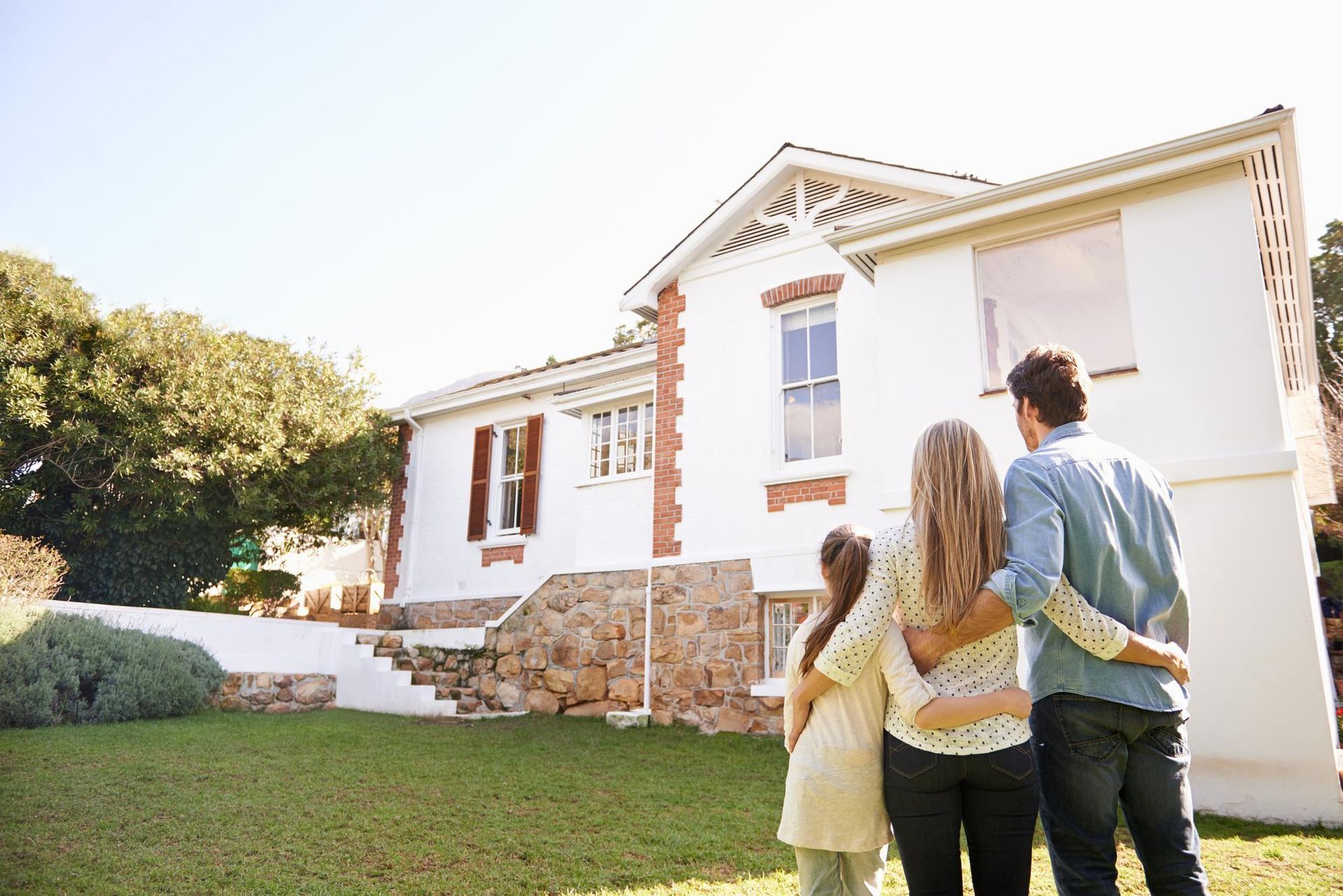 Three people stand on a lawn facing a white house with stone foundation in bright sunlight