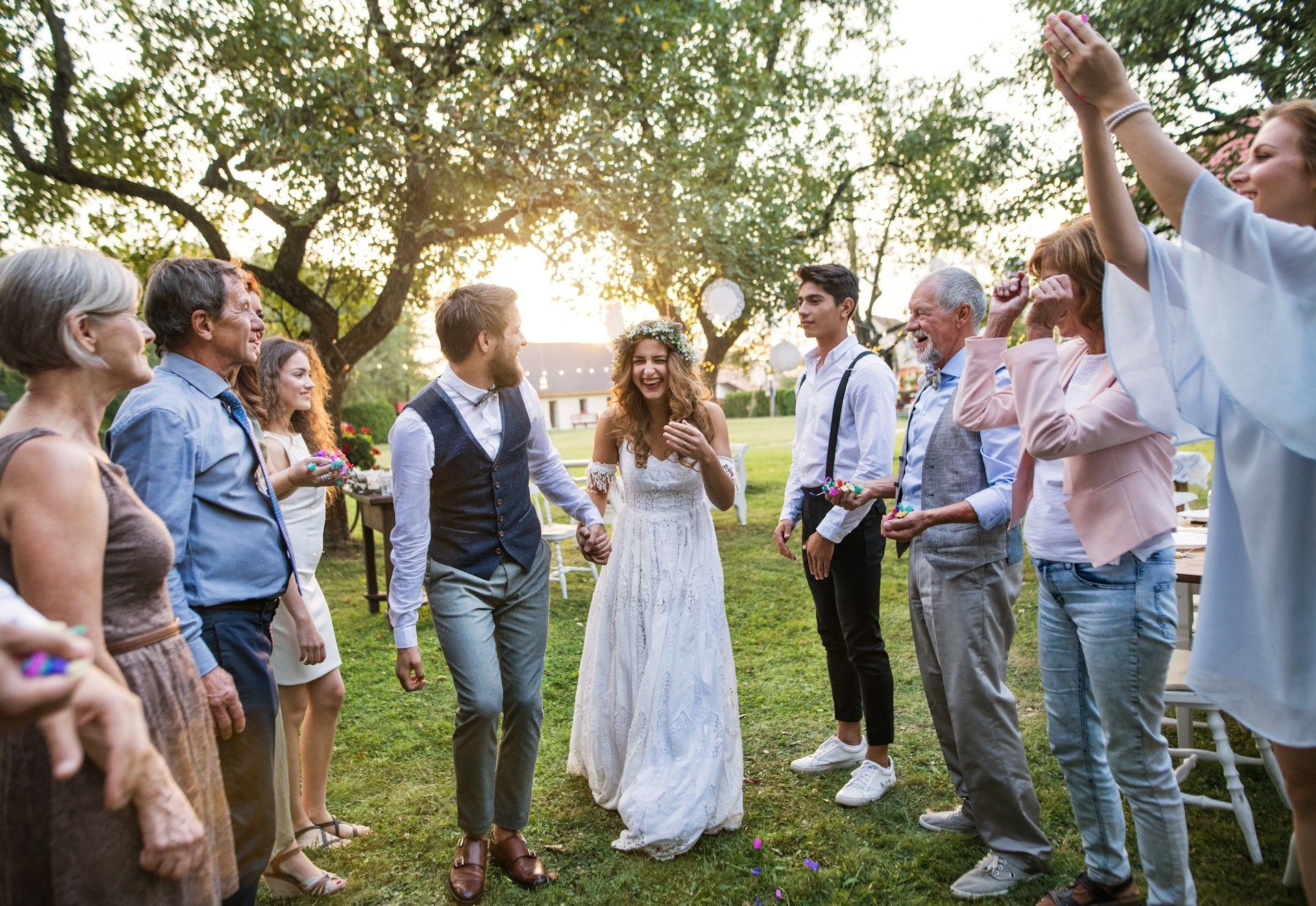 Wedding guests dancing outdoors under trees at sunset, with the bride and groom smiling in the center