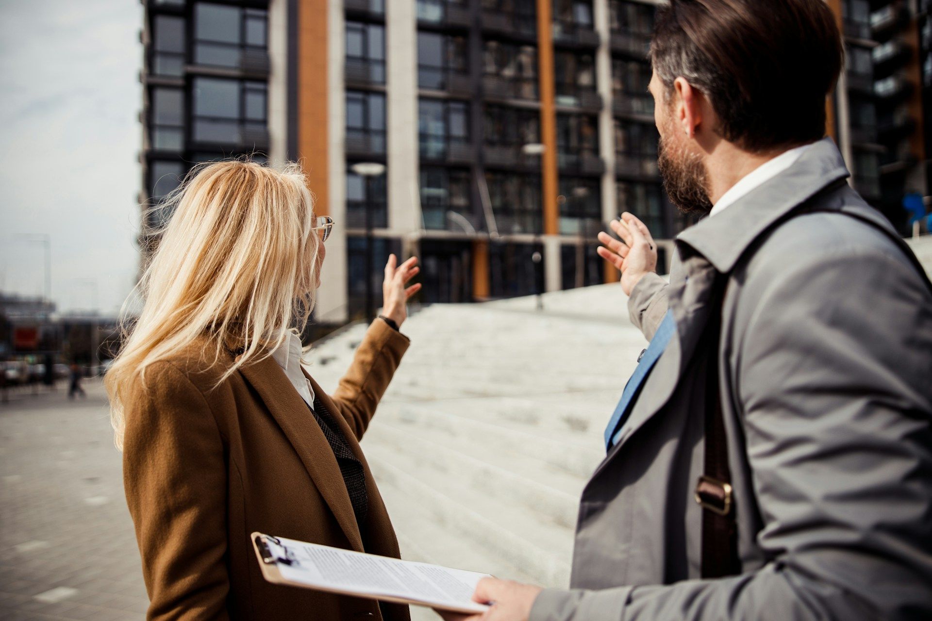 Three people in coats discussing a tall office building outdoors, one holding a clipboard