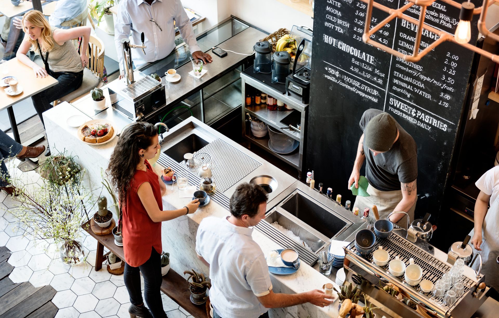 People at a café counter ordering drinks, with a chalkboard menu and pastry display.
