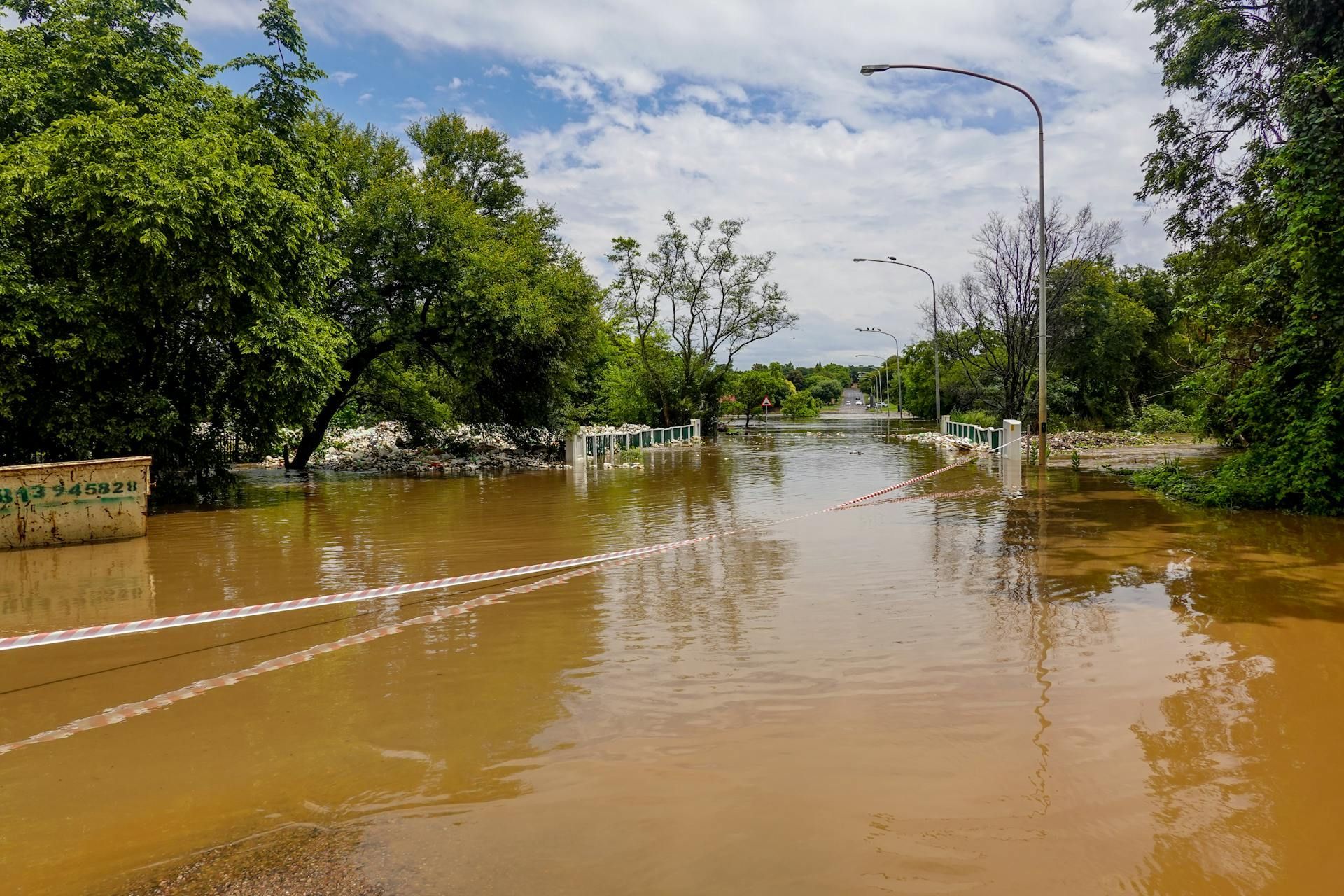 Flooded tree-lined road with muddy water and streetlights under a partly cloudy sky