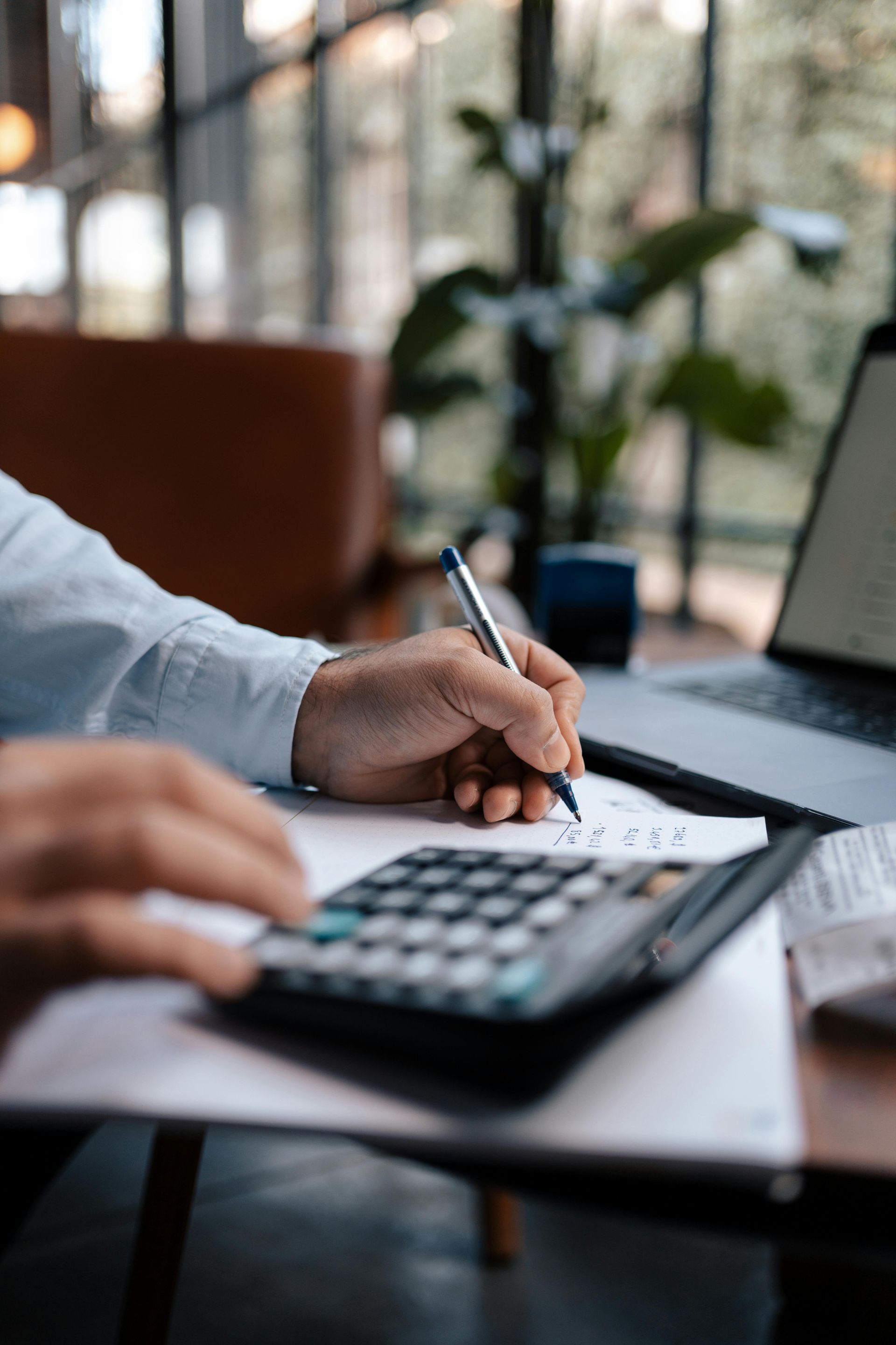 Hands calculating with a calculator while writing notes beside a laptop in a bright office
