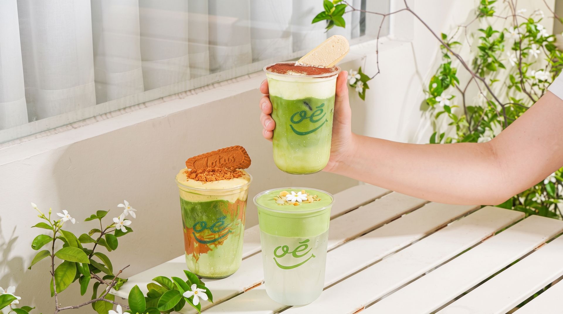 Hand holding a green layered drink on a white table with two matching cups and potted plants nearby