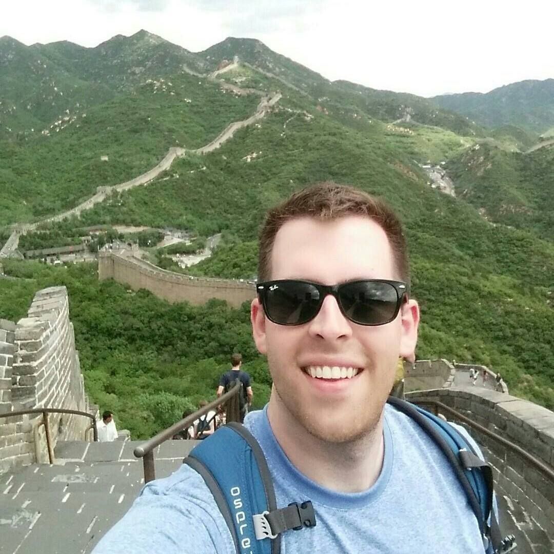 Brian Voss smiling in sunglasses on the Great Wall of China with green mountains behind him